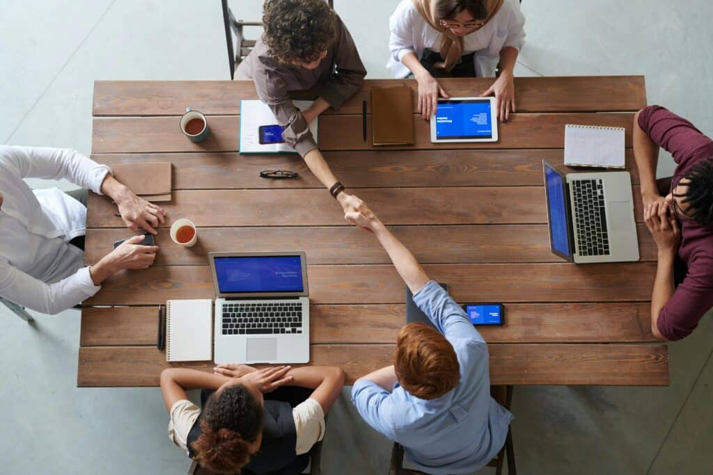 Vier Personen vor Laptops sitzen um einen Tisch. Zwei Personen reichen sich die Hände zum Handschlag als Zeichen des Einverständnisses.