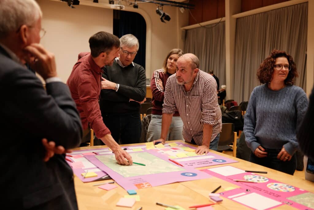 Sechs Personen stehen um einen Tisch, auf dem ein Projekt-Canvas liegt. Ein Teilnehmer zeigt dem Moderator, wo er seine Ressourcen einbringen kann.